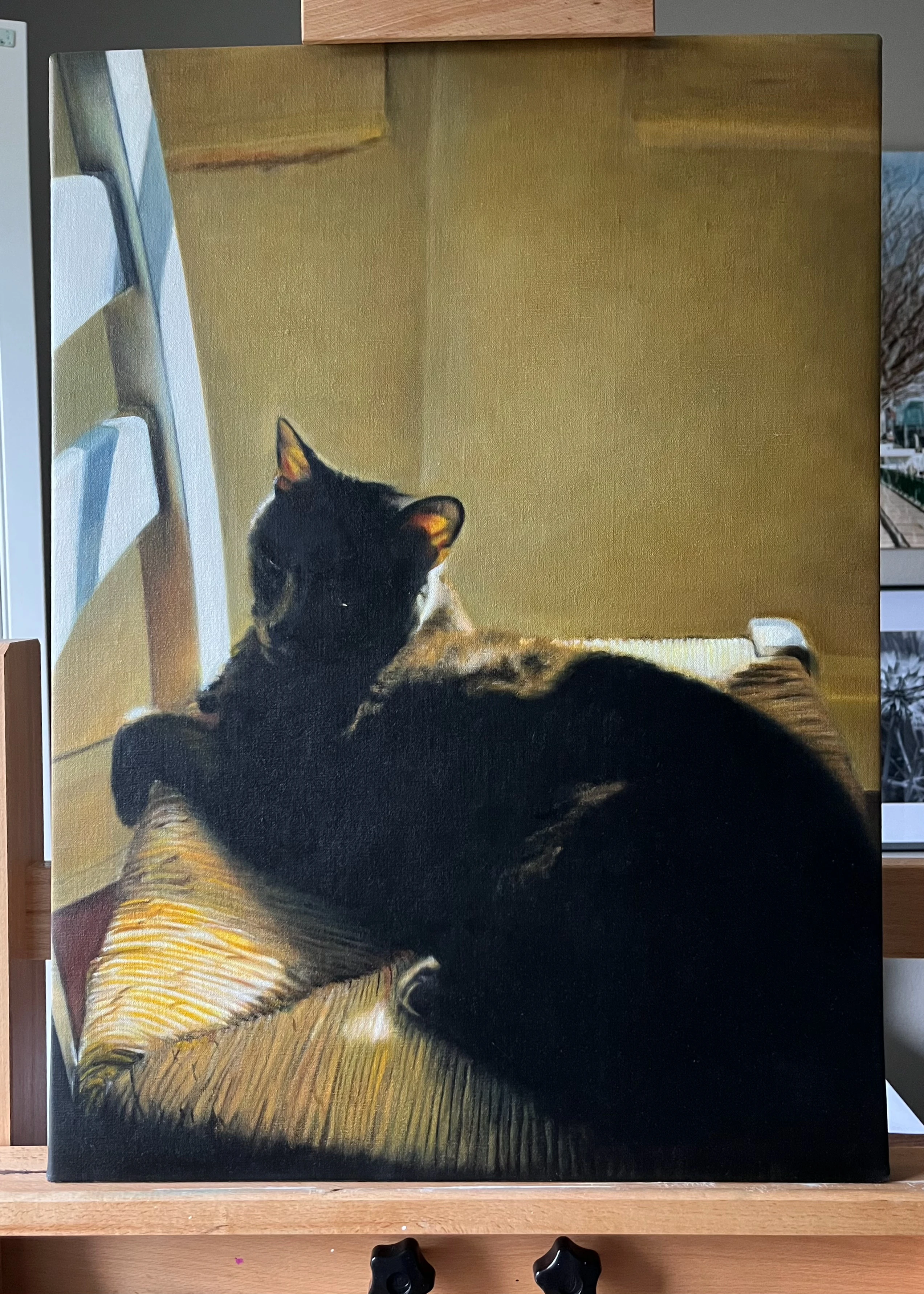stretched oil painting, sitting on a wooden easel, of a dark grey cat lounging on a wicker dining chair