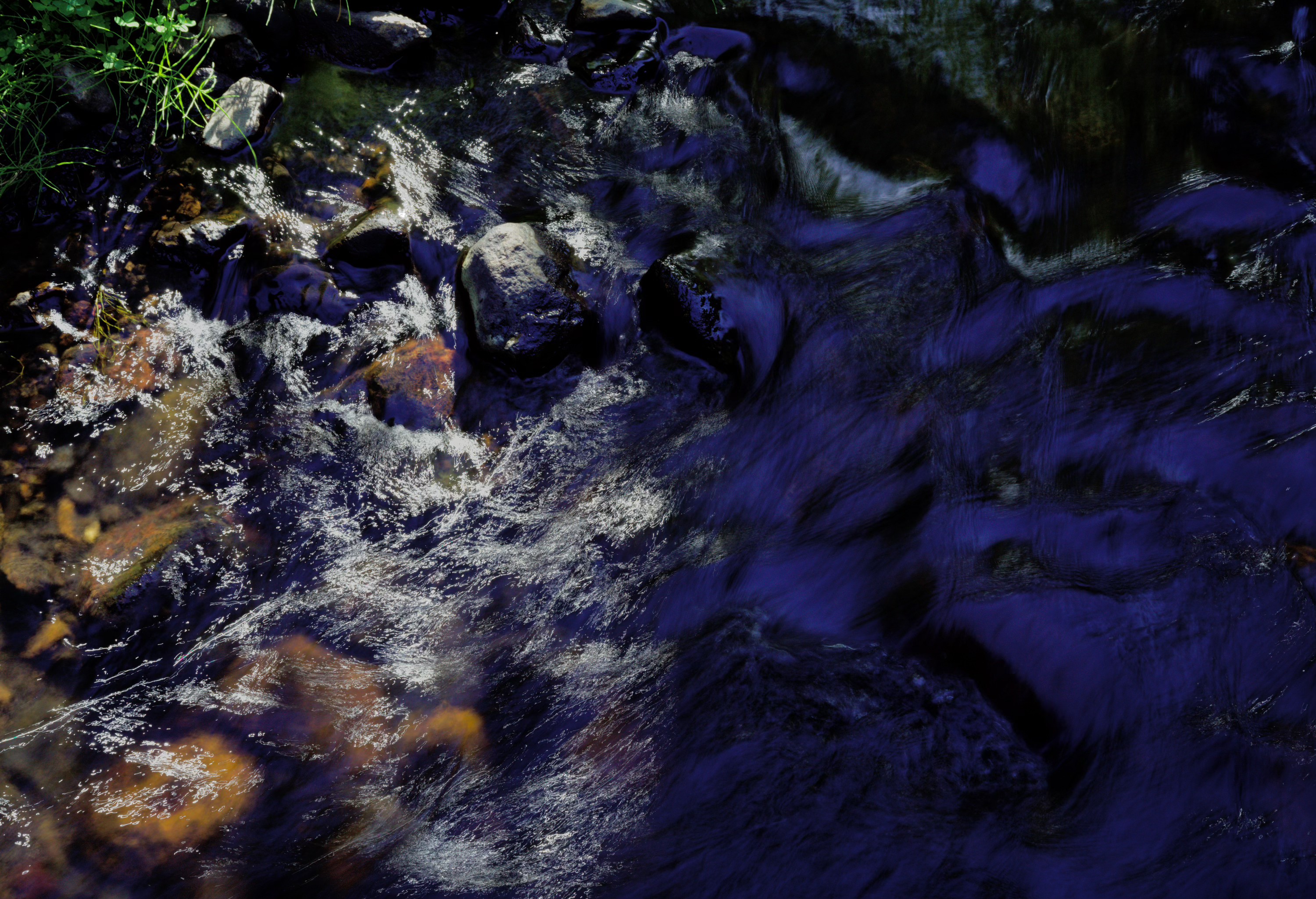 photograph of a running stream, split down the middle: the left is covered with traces of sun reflecting in the moving water, while the right is shadowed, picking up splotchy dark reflections of the sky
