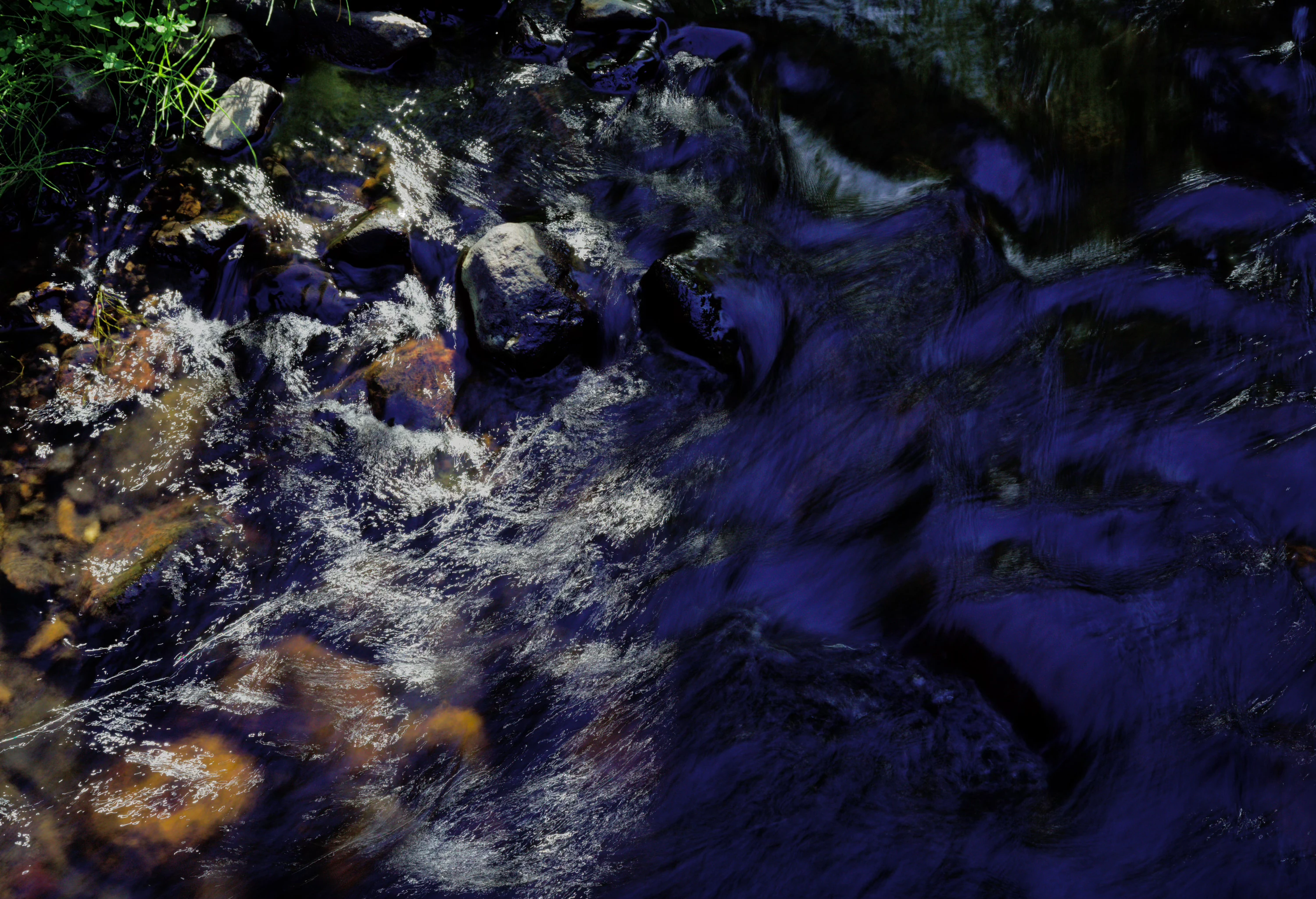 photograph of a running stream, split down the middle: the left is covered with traces of sun reflecting in the moving water, while the right is shadowed, picking up splotchy dark reflections of the sky