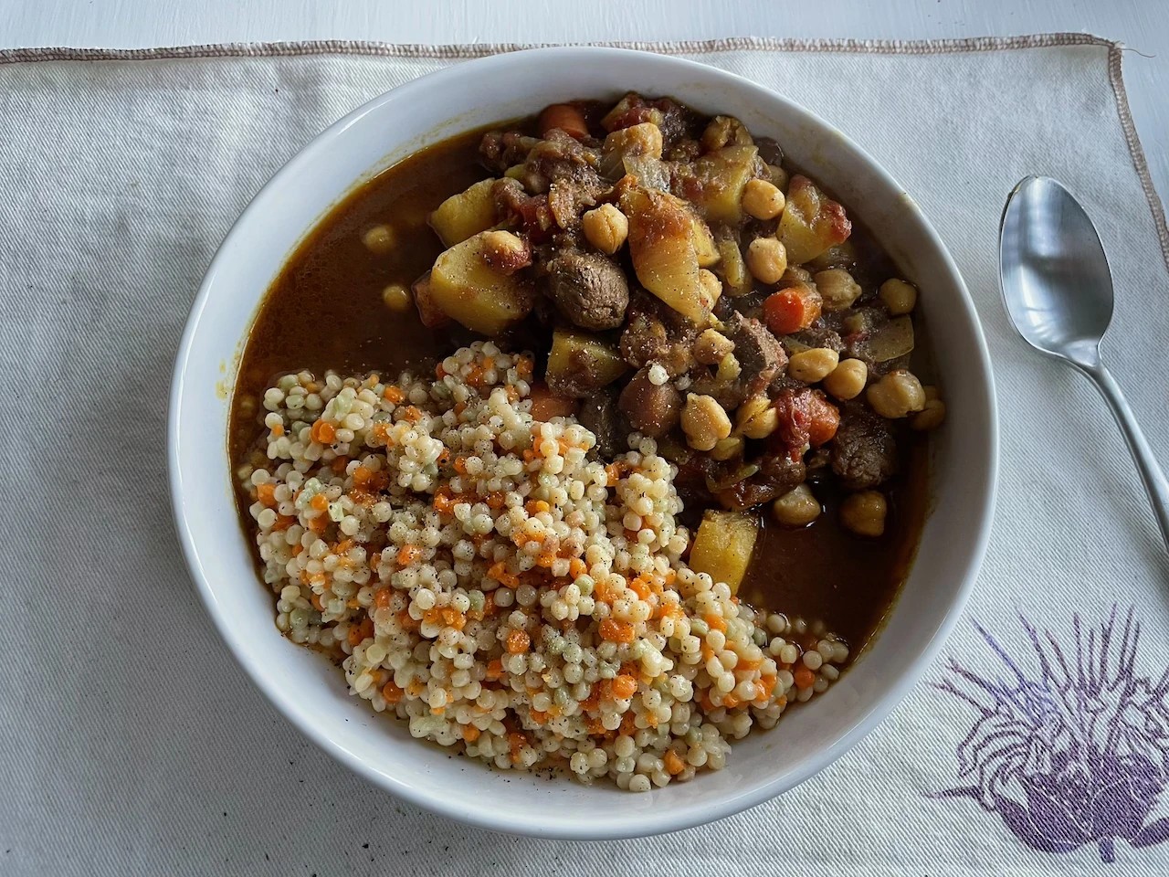 A white bowl of Moroccan lamb stew with chickpeas, carrots, and potatoes, served with pearl couscous