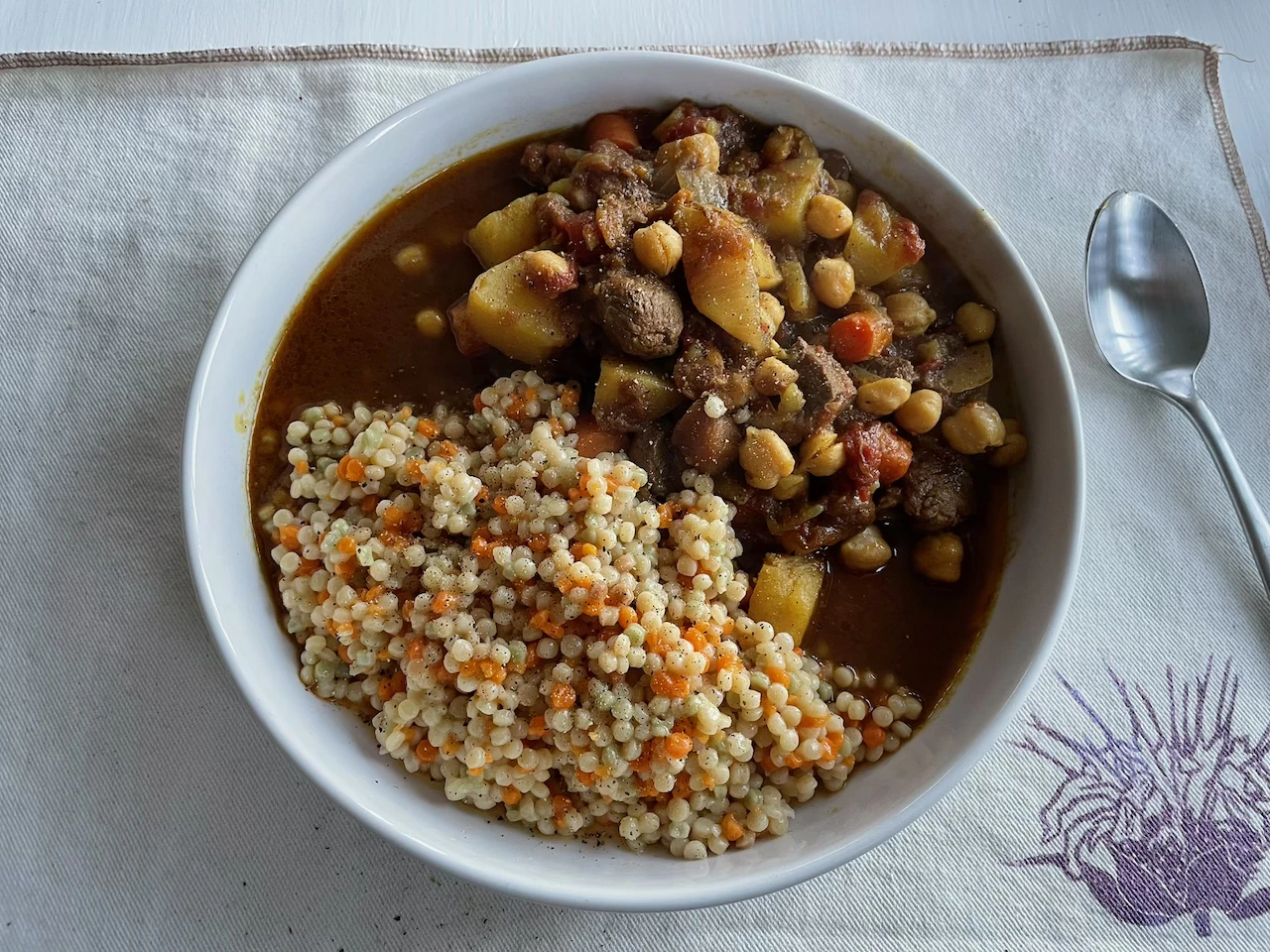 A white bowl of Moroccan lamb stew with chickpeas, carrots, and potatoes, served with pearl couscous